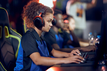 Side view of a focused mixed race girl, female cybersport gamer wearing headphones playing online video games, participating in eSport tournament