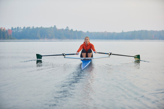 Middle aged woman rowing on calm autumn lake at early morning with mist over water.