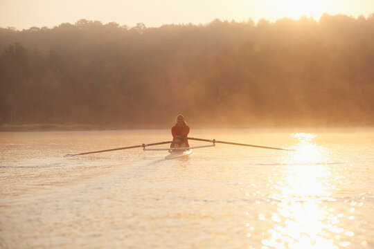 Middle aged woman rowing on calm autumn lake at early morning with mist over water.
