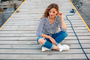 A woman in jeans and a shirt sits on the pier and looks at the yachts. Girl on the shore near the ships and boats.