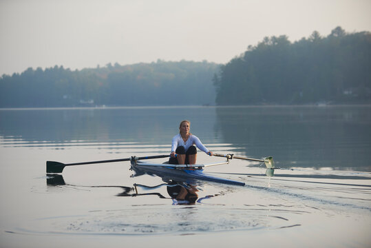 Middle aged woman rowing on calm autumn lake at early morning with mist over water.