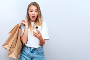 Young female in casual cloth with paper shopper bags holding and looking to smartphone with shocked expressions on grey backdrop. Shopping. Black friday and cyber monday sales. Online shopping