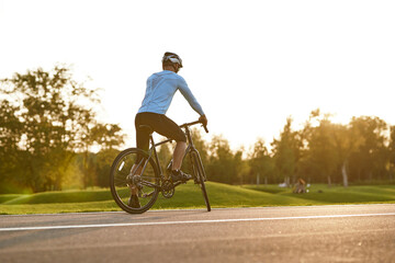 Rear view of athletic man in sportswear cycling outdoors at sunset, professional racer standing with bicycle on the road and enjoying amazing nature view