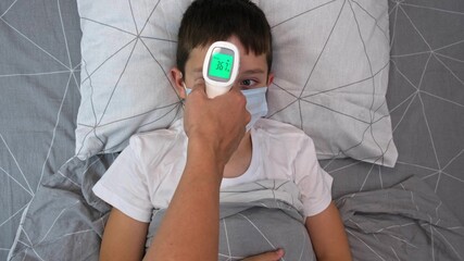 A recovering boy medical protective mask lying in bed and his mother measuring temperature with infrared contactless thermometer, concept of healthy child