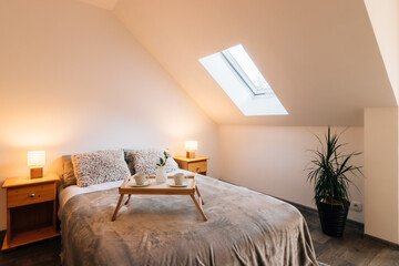 Small attic bedroom in a family house with morning breakfast service on the bed. The bed also has a fluffy blanket and two fluffy pillows, on the sides there are bedside tables with lit lamps.