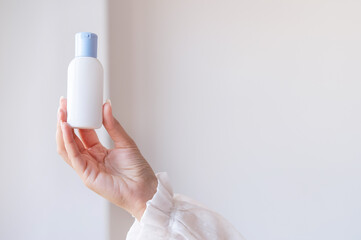 Woman in white blouse holding white antiseptic sanitizer bottle on hand against white wall. Hand hygiene, antibacterial protection. Unrecognized personality