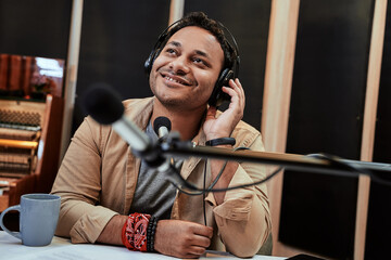 Happy young male radio host looking aside while broadcasting in studio, using microphone and headphones