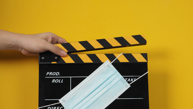 Hand Is Holding Black And Yellow Color Clapper Board Or Movie Slate With Face Mask. It Use In Video Production And Cinema Industry On Yellow Background.