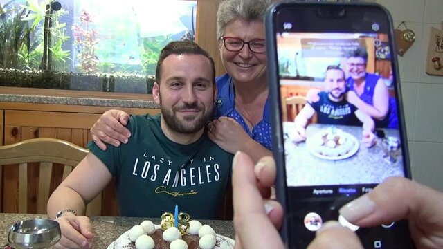 Young man celebrating his birthday with his mother kissing him while another person takes photos with her smartphone.