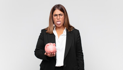 young woman with a piggy bank feeling disgusted and irritated, sticking tongue out, disliking...