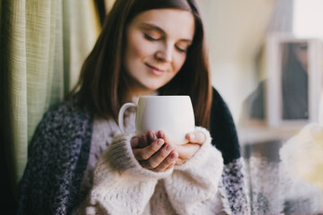 Young woman in white knitted sweater drinking tea near window