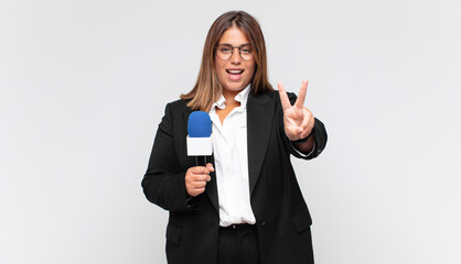 young woman reporter smiling and looking happy, carefree and positive, gesturing victory or peace...