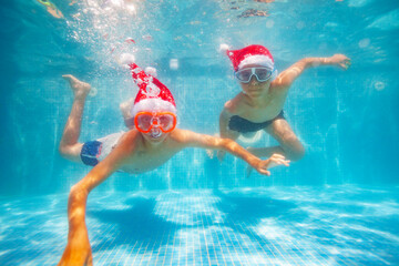 Obraz na płótnie Canvas Two cool boys in scuba masks dive and swim underwater wearing Santa Claus hat in the pool