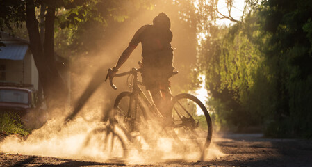 Cyclist riding a bicycle on a gravel road at sunset. A silhouette of young sporty man on a gravel bike in a cloud of dust from real whell after skidding.