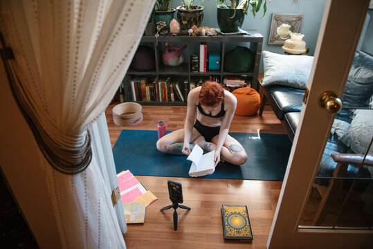 Woman With Tattoos Reading Yoga Book On Mat At Home