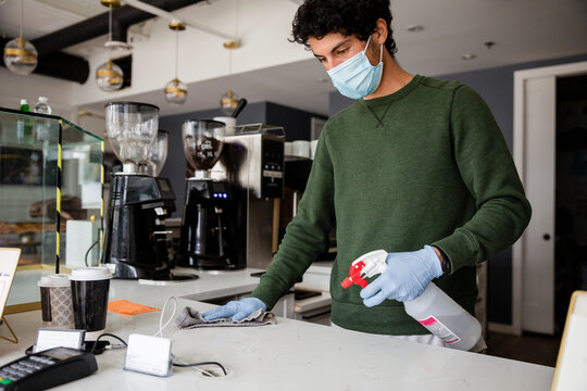 Young male barista in face mask and gloves disinfecting cafe counter