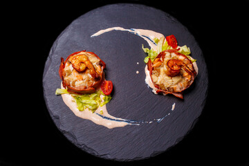 Close-up of medallions with tuna, salmon, chicken, bacon, greenery, cherry tomatoes and prawns in sauce on the dark round tray isolated on the black background. Top view