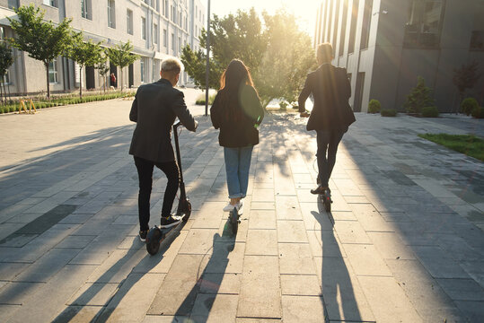 Back view of joyful international friends holding electric scooters going near modern building and talking.