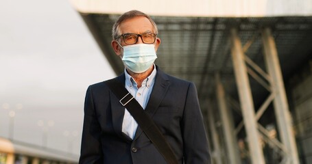 Portrait shot of handsome Caucasian old man in glasses and medical mask standing outdoors in urban environment and looking at camera. Gray-haired senior male in eyeglasses and respiratory protection.