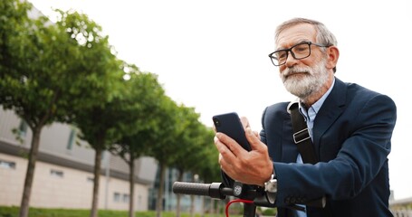 Senior gray-haired man in glasses and headphones standing at bike on street and tapping or scrolling on mobile phone. Old grandfather in eyeglasses using smartphone and browsing online outdoor.