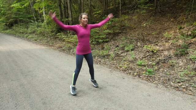 Athletic 40s woman doing jumping jacks on forest trail. Warming up.
