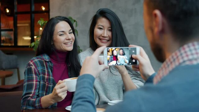 Guy is taking pictures of women friends posing with coffee cups in cozy cafe using smartphone camera. Friendship and contemporary devices concept.