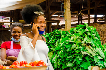 young pretty african businesswoman standing in the market making a phone call wearing face mask to prevent herself from the outbreak in the society.