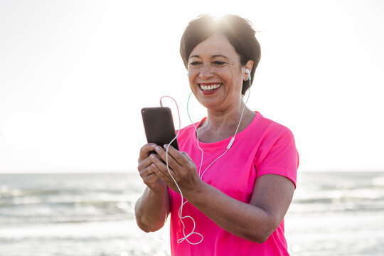 Happy Senior Woman Tracking Work Out Fitness From The Phone Outdoor. 
Sixsties Woman Wearing Headphones Doing Jogging At The Beach. Concept About Mature People, Sport And Technology. 