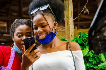 young beautiful african trader and her customer wearing face mask feeling excited about on their cellphone