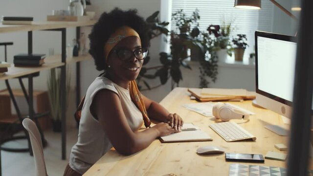 Young beautiful African American businesswoman using computer at desk in the office, then turning to camera and posing with smile while working late in the evening