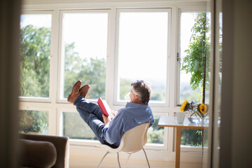 Carefree senior man reading book with feet up at sunny window