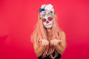 young beautiful woman wearing halloween make up over red background holding something with open palms, offering to the camera.