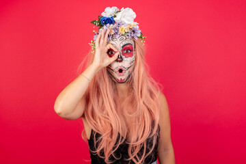 young beautiful woman wearing halloween make up over red background doing ok gesture shocked with surprised face, eye looking through fingers. Unbelieving expression.