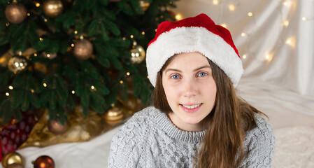 Portrait of a young woman in a gray sweater and santa claus hat sits near a Christmas tree against the background of a glowing garland.