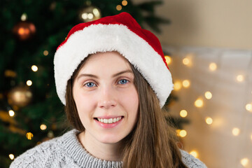 Portrait of a young woman in a gray sweater and santa claus hat sits near a Christmas tree against the background of a glowing garland.