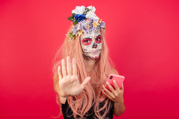 Young beautiful woman wearing halloween make up over red background using smartphone with open hand doing stop sign with serious and confident expression, defense gesture