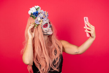Young beautiful woman wearing halloween make up over red background, smiling and taking a selfie ready to post it on her social media.