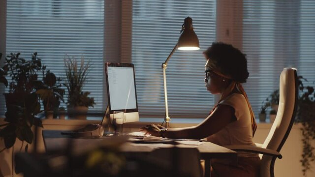 Side view of concentrated afro-american businesswoman sitting at office desk and working on computer in the evening