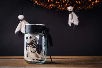 Funny Halloween day decoration party, The white ghost scary face and black spider in jar glass on wooden table, studio shot isolated on black background, Happy holiday concept