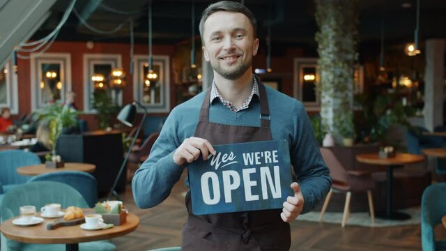 Slow motion portrait of confident young man in apron business owner holding open sign in cafe smiling and looking at camera. People and start-up concept.