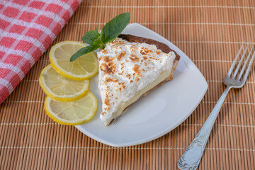 A slice of homemade lemon meringue pie on a plate, with slices of lemons, napkin completing the scene in a rustic setting and wooden planks background