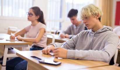 Portrait of positive teenage male pupil sitting at desk studying in classroom