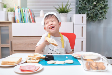 Cute Asian boy wearing chef hat and apron having fun preparing sandwiches, Little kid holding bread while making sandwiches at home