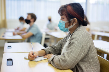 Diligent asian teen girl in protective mask studying in college with classmates. New life reality...