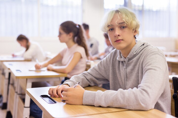 Portrait of positive teenage male pupil sitting at desk studying in classroom