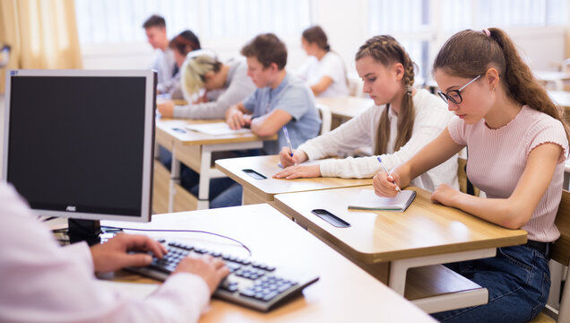 Focused teenage students studying in classroom with teacher, writing lectures in workbooks