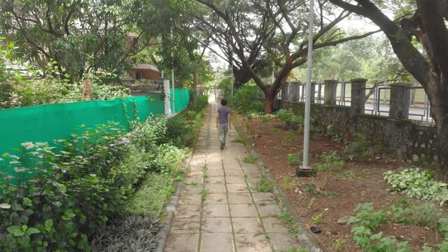 high pan follow shot of a young tan caucasian man black hair walking down a shaded paved overgrown tree path