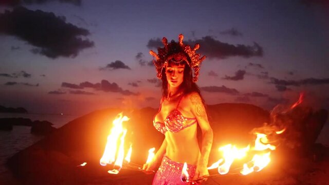 Erotic and sensual belly dancer performing spinning and figure 8 moves with fire props on the beach during golden hour.