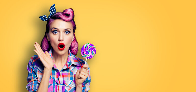 Purple head excited very surprised woman with lollipop. Pin up girl with wide opened mouth, eyes. Beauty model at retro fashion and vintage studio concept Yellow orange color background with copyspace