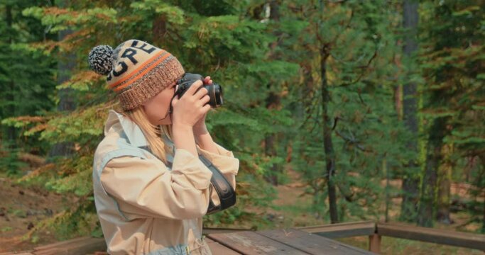 Photographer woman taking picture of fall forest on camera. Slow motion of beautiful smiling traveler in scenic green woods. Young female exploring nature with pine trees on motion background, 4K USA